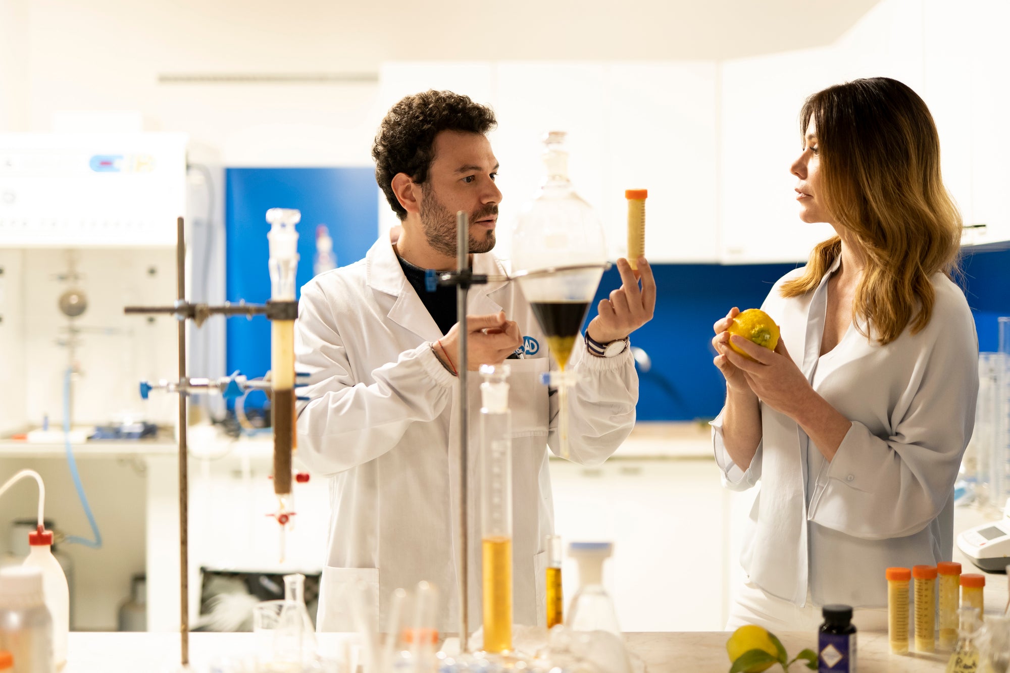A scientists in a laboratory setting, one holding a beaker and Naomi holding a citrus bergamot fruit, with lab equipment around.